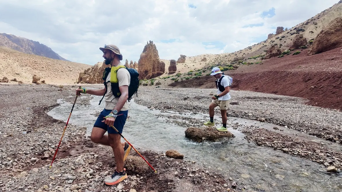 Deux coureurs d'ultra-trail traversent une rivière à gué dans une vallée minérale de l'Atlas marocain, sur le Trans Atlas Marathon.